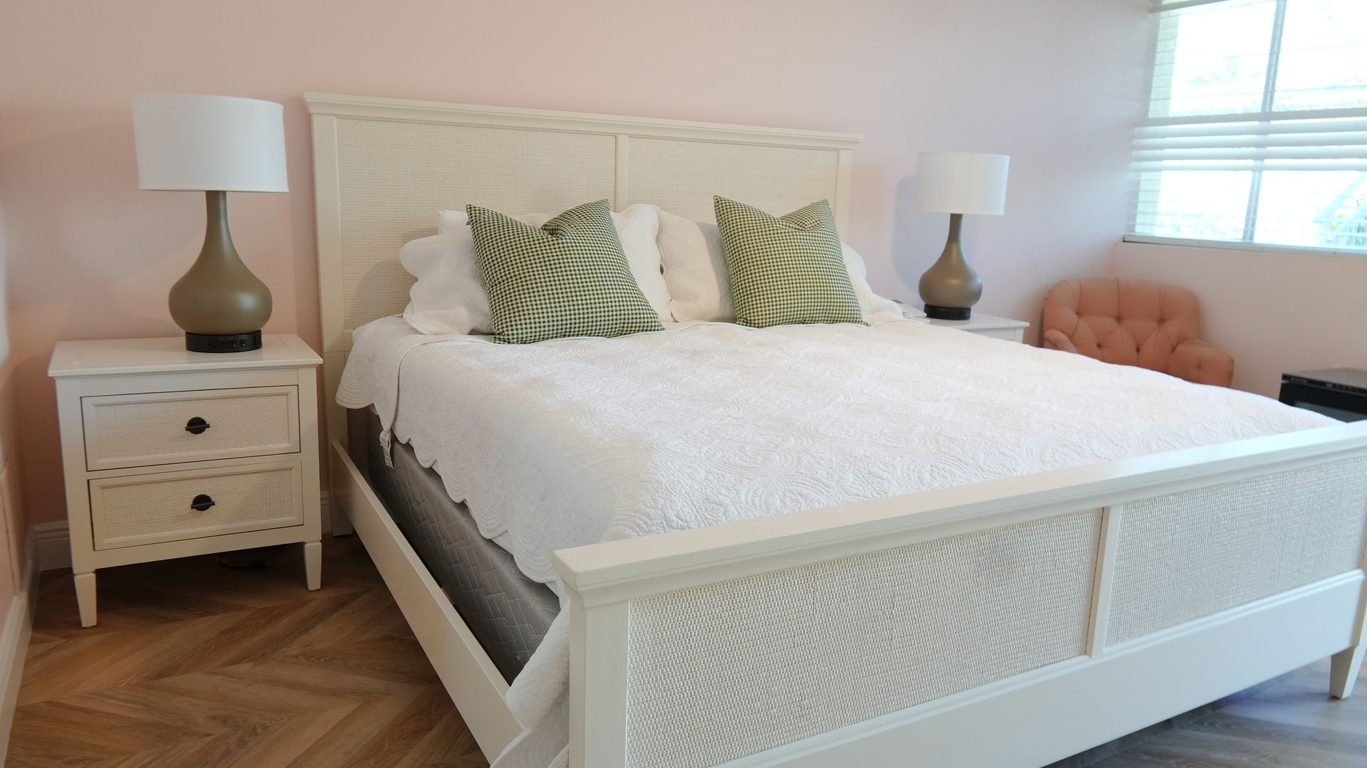 Bedroom with white bed frame, nightstands, lamps, and pink walls. Two green patterned pillows on the bed.