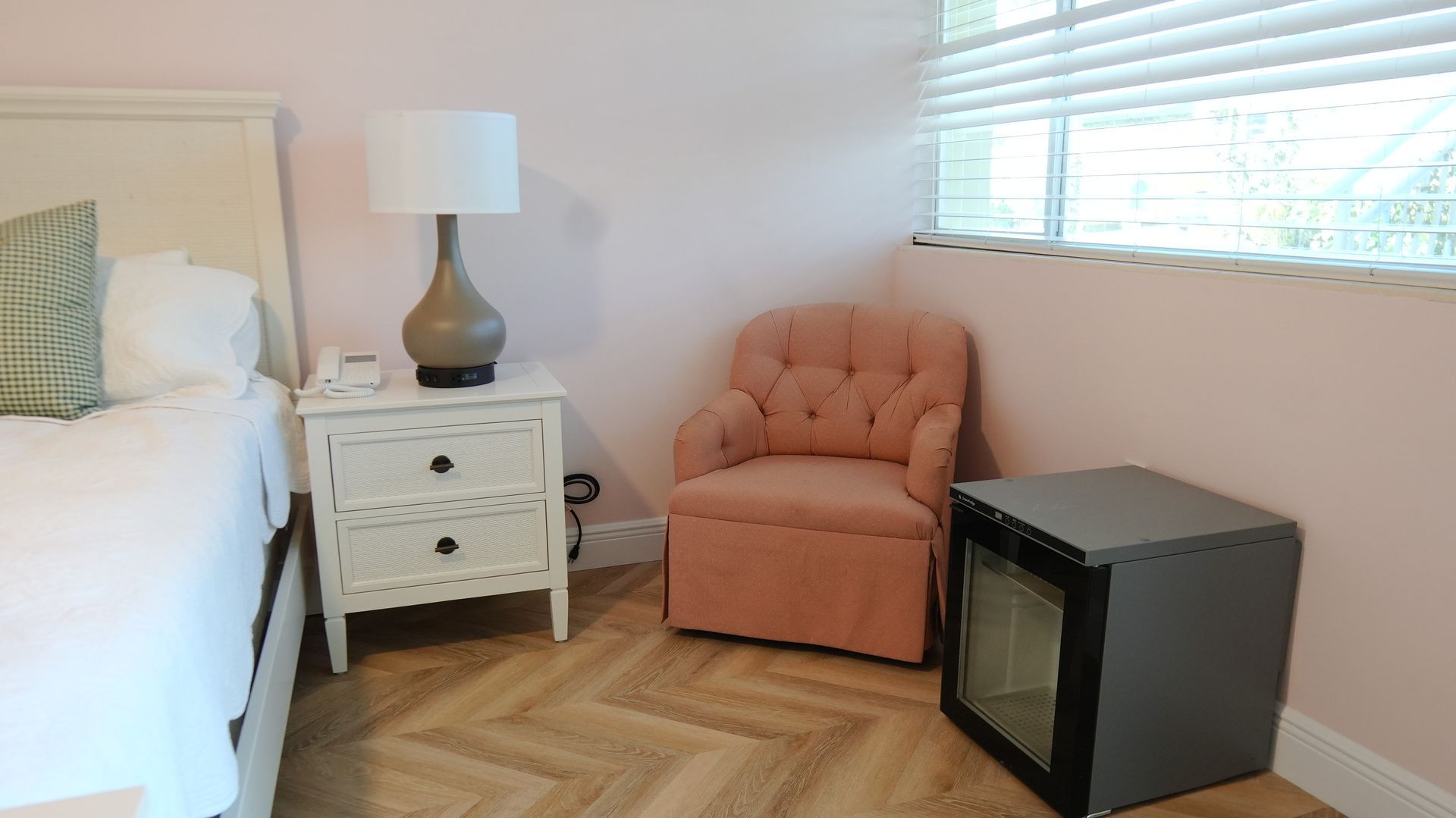 Bedroom corner: white bed, nightstand with lamp, pink chair, small fridge, and window.