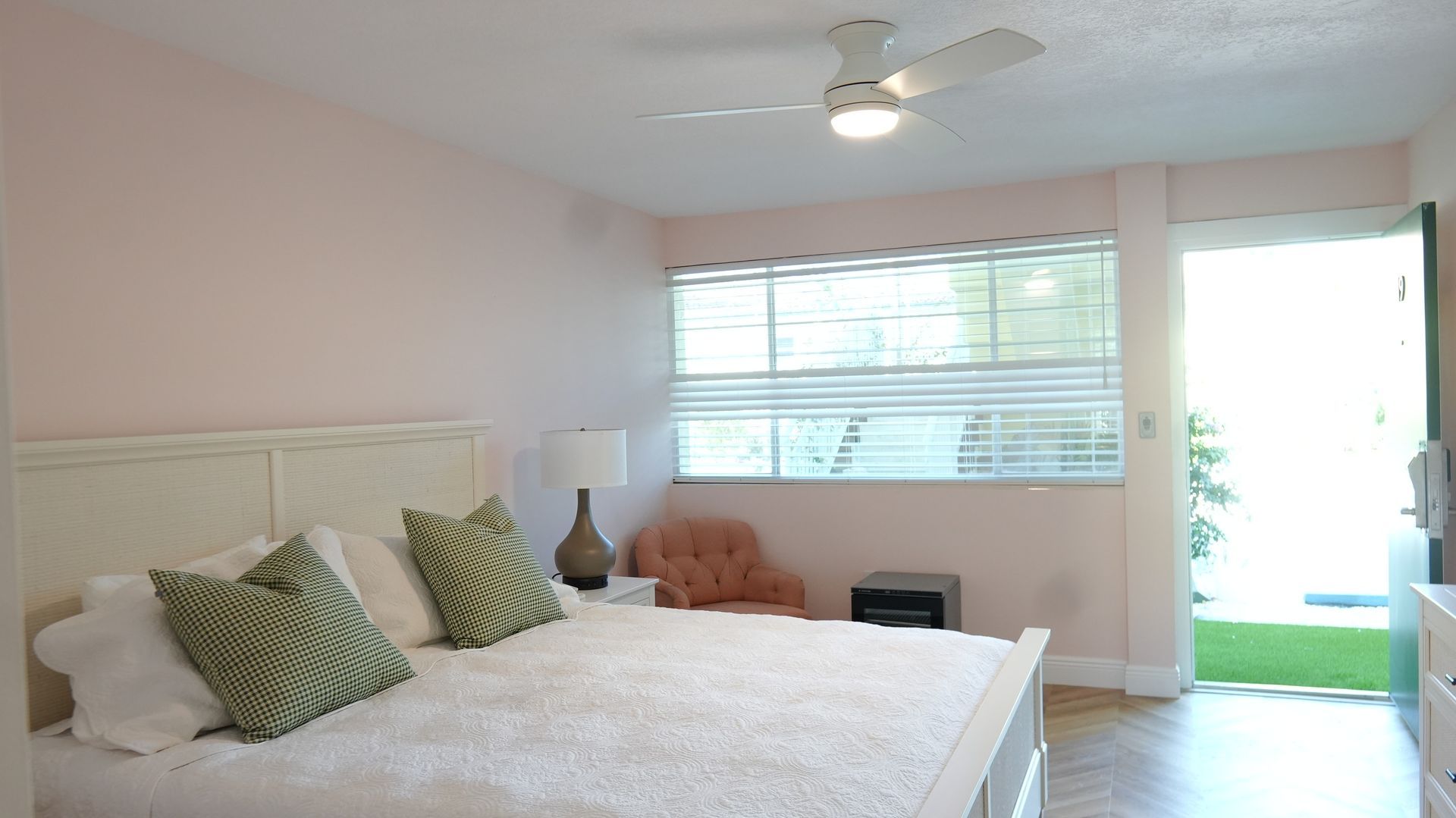 Bedroom with a white bed, pink walls, a window with blinds, and an open doorway leading outside.