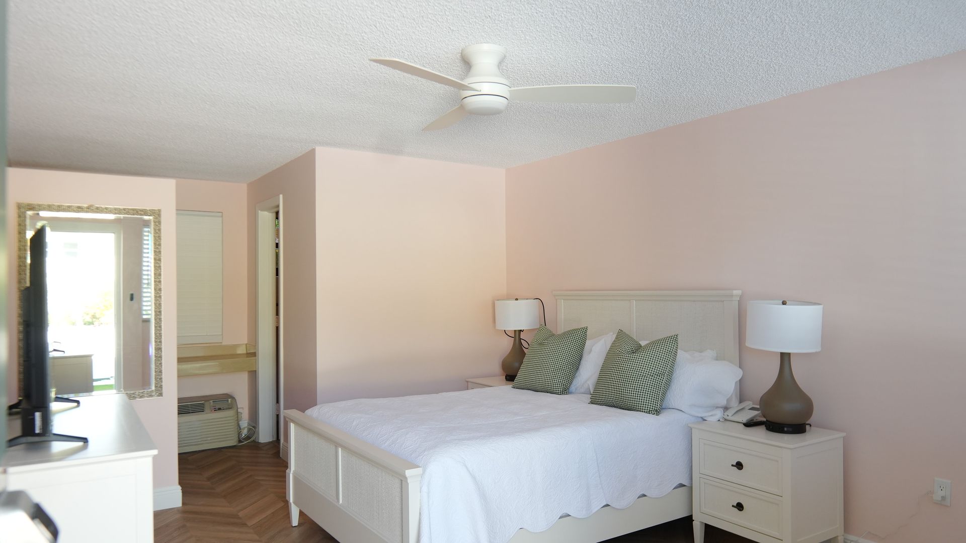 A bedroom with a white bed, pink walls, and a ceiling fan. A TV and a mirror are visible.
