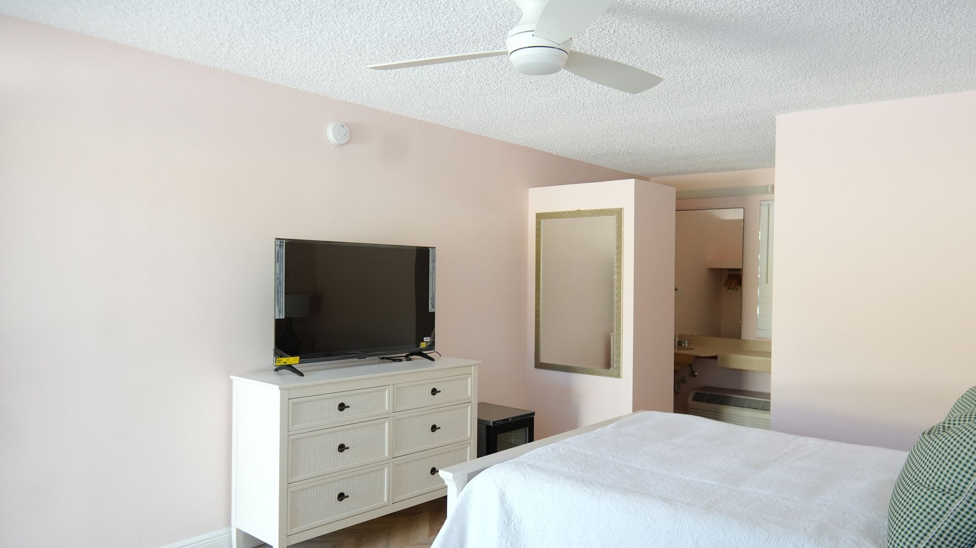 A light pink bedroom with a white dresser, TV, and ceiling fan; a bathroom is visible in the background.