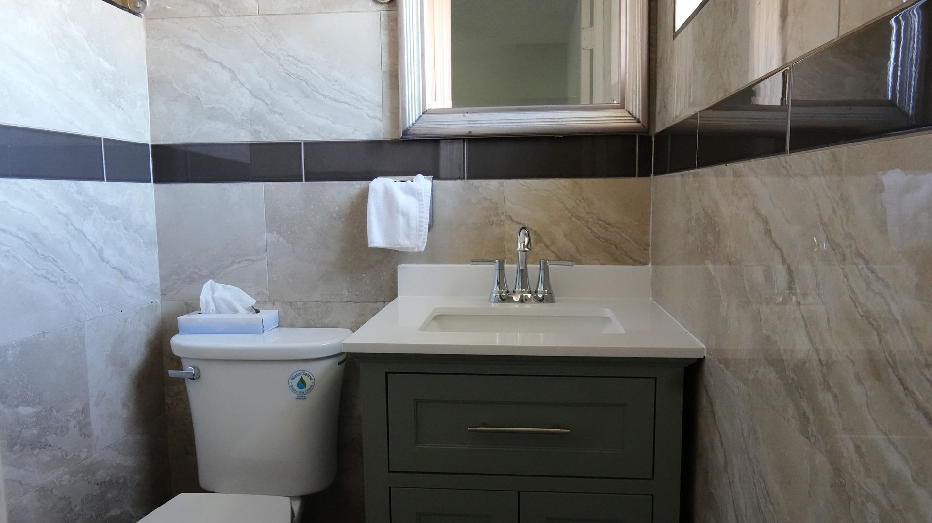 Bathroom with a toilet, sink, and cabinet. Gray and beige tile walls. White countertop.