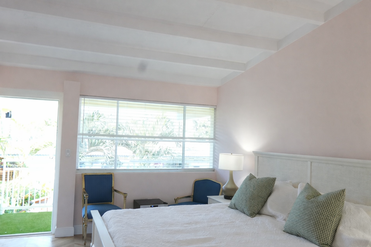 Bedroom with a white bed, pink walls, open doorway, and a window with blue chairs.