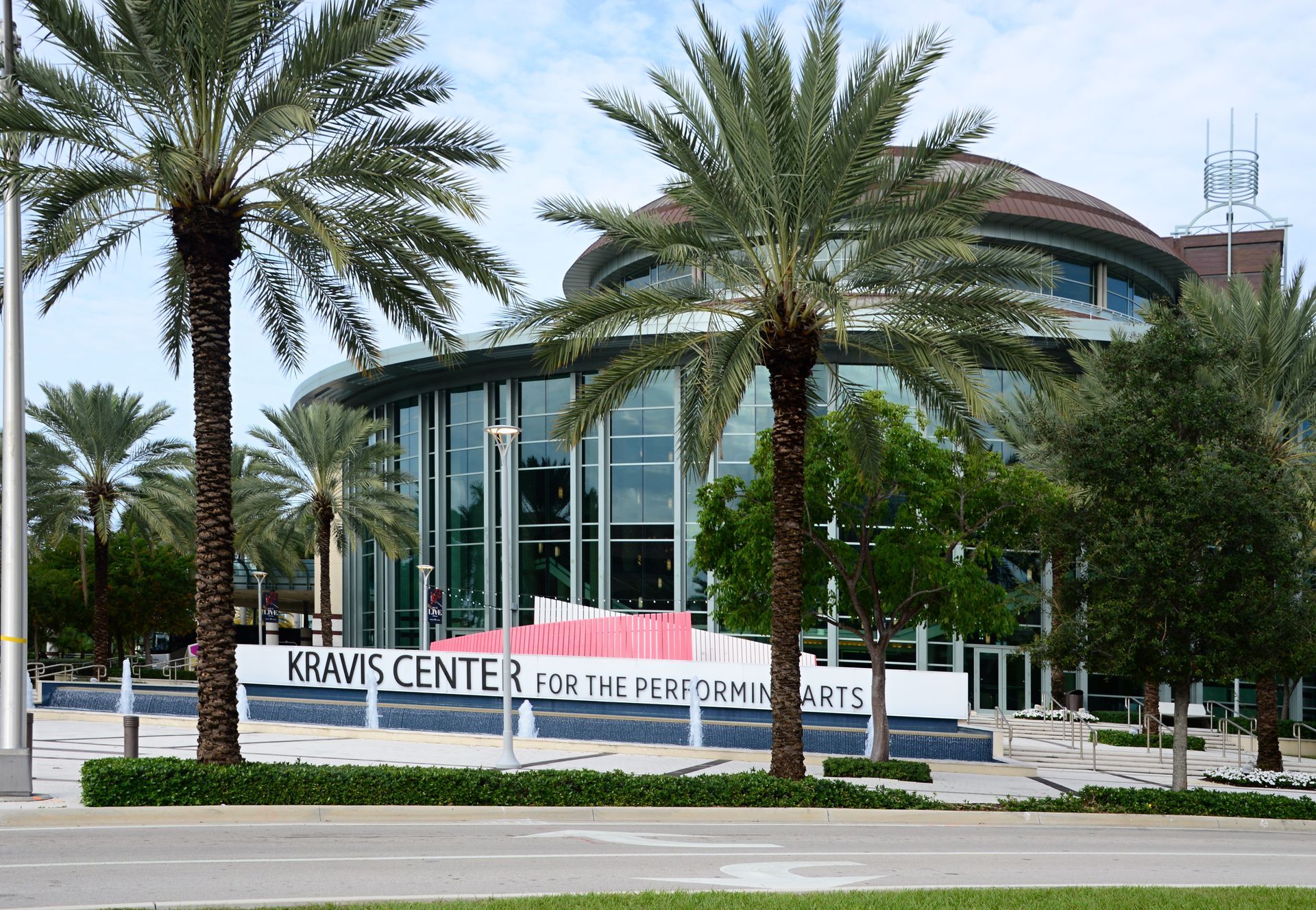 Exterior of the Kravis Center for the Performing Arts with palm trees and a sign in front of a modern glass building.