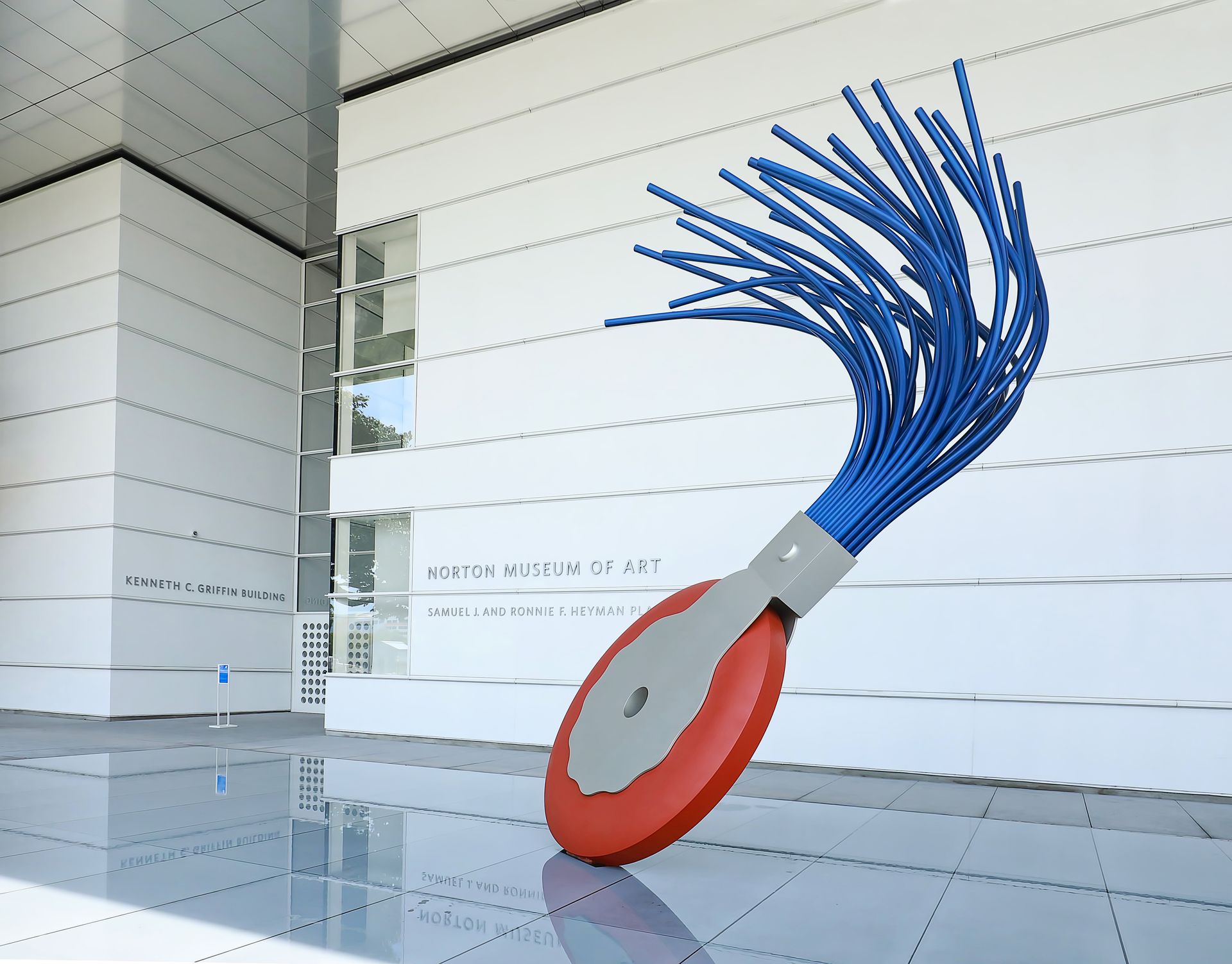 Sculpture of a red, grey, and blue brush against a white building.