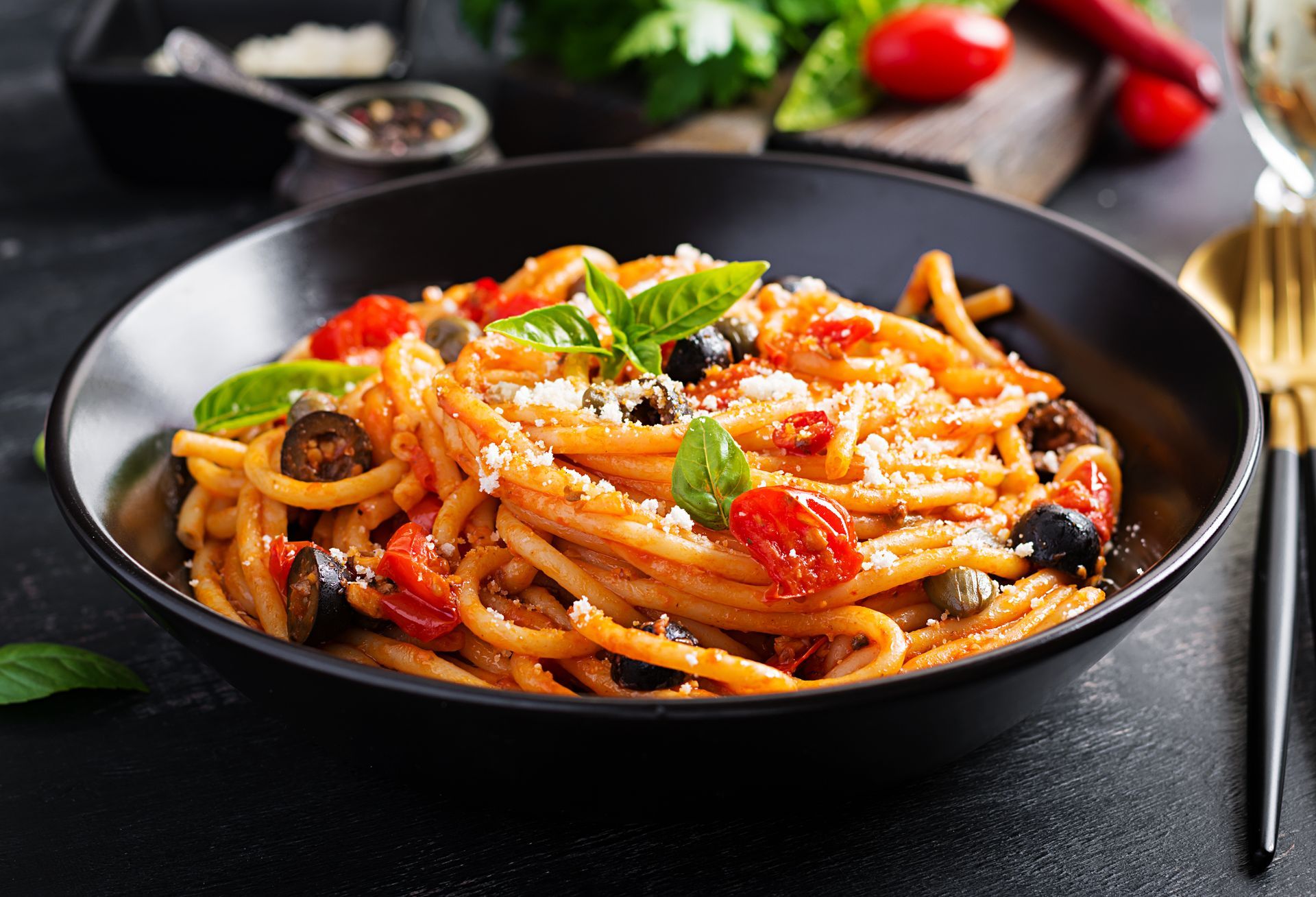 Pasta dish with tomatoes, olives, capers, basil, and cheese in a black bowl.