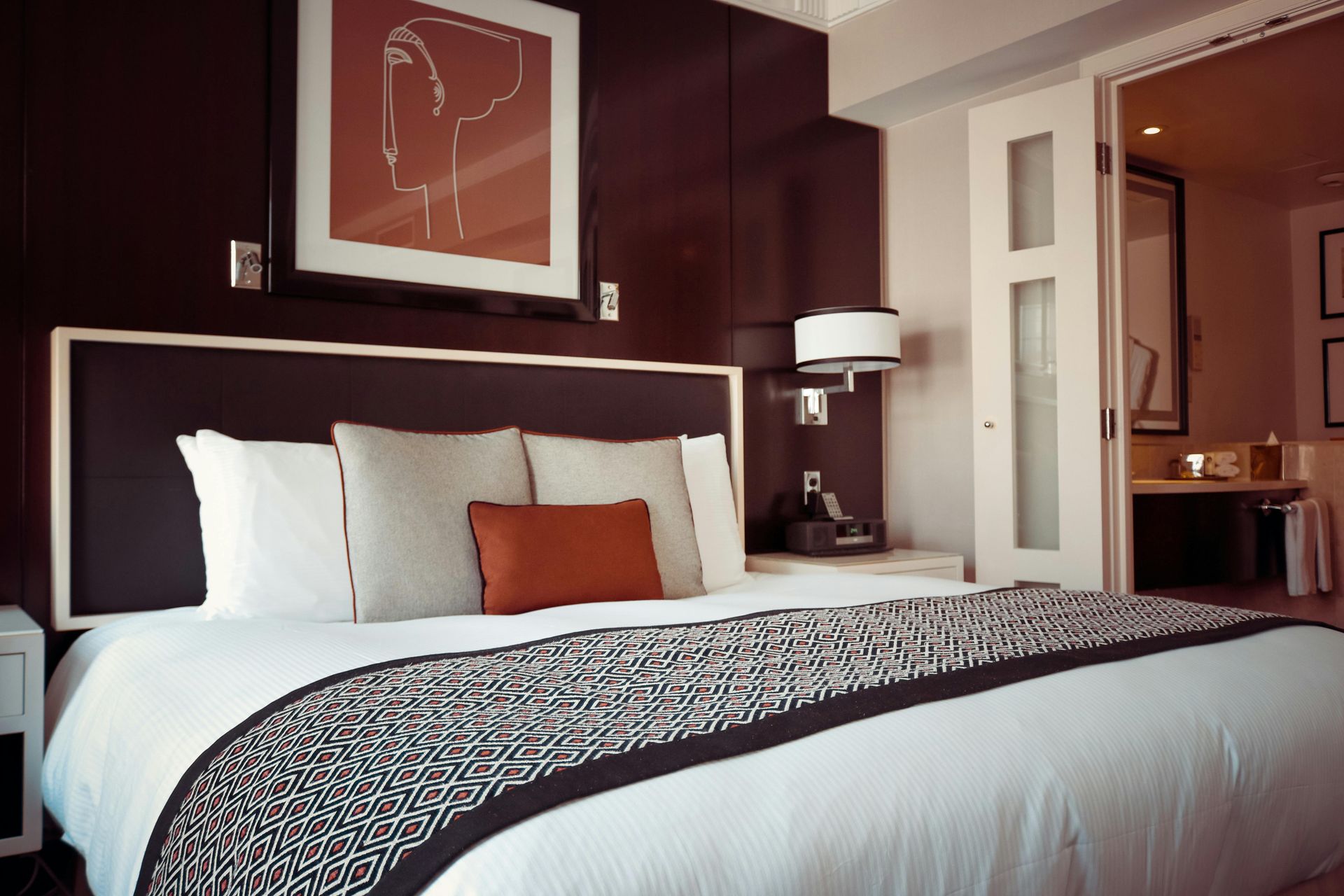 Hotel bedroom: Large bed with white linens and patterned runner, dark headboard, framed artwork, and bathroom access.