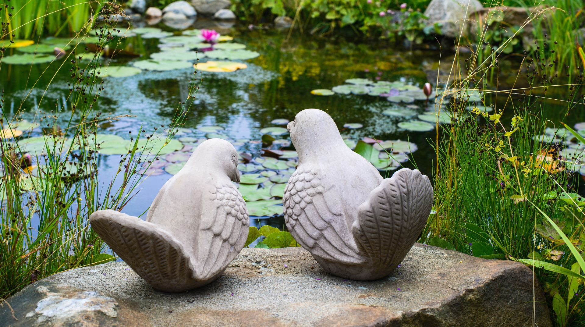Two stone bird statues sit perched on a rock overlooking a garden pond filled with lily pads.