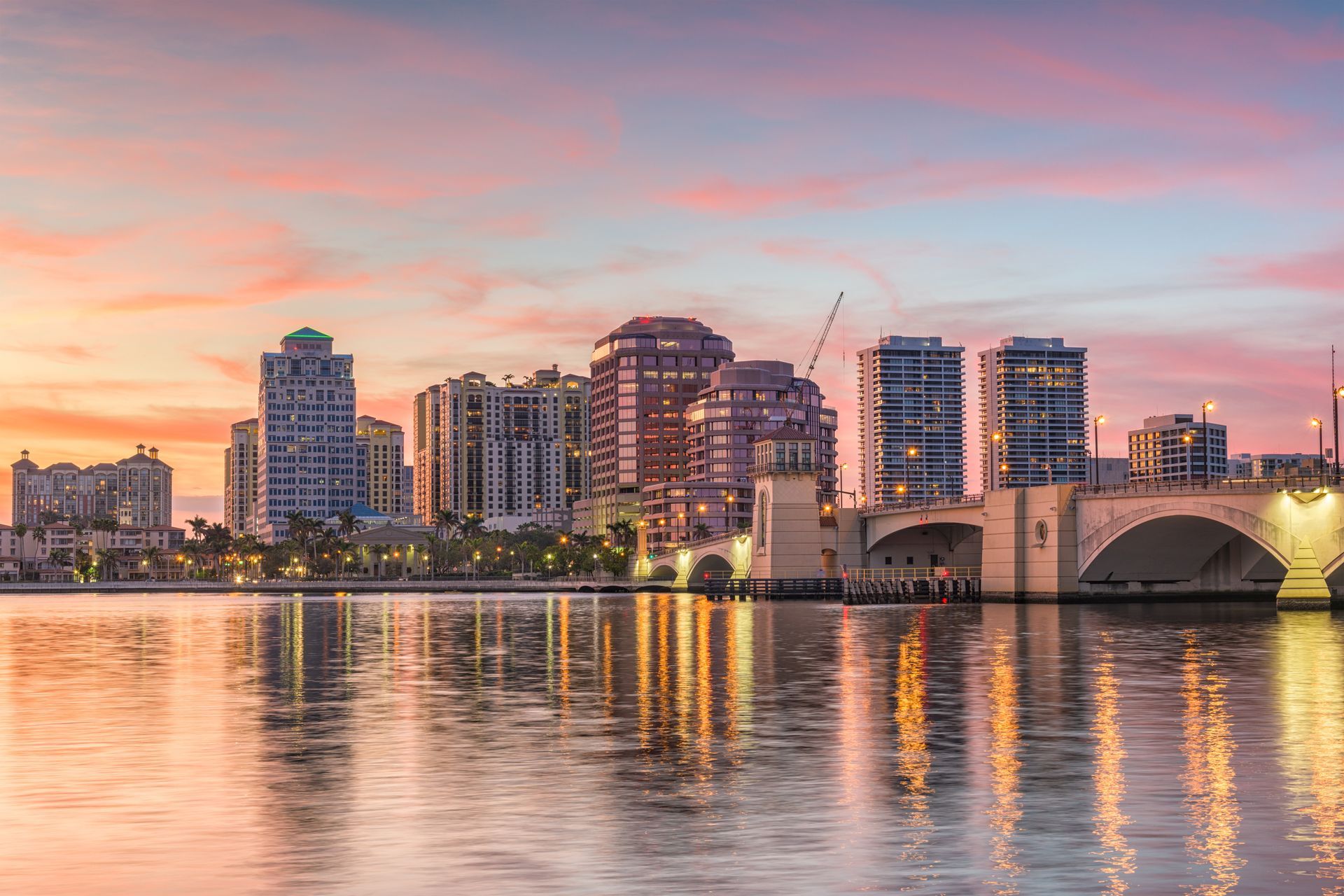 West Palm Beach skyline and bridge reflected in the water at sunset with pink and orange hues.