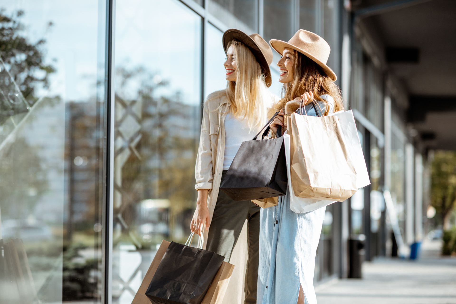 Two women with shopping bags walking outside a storefront in hats and light clothes