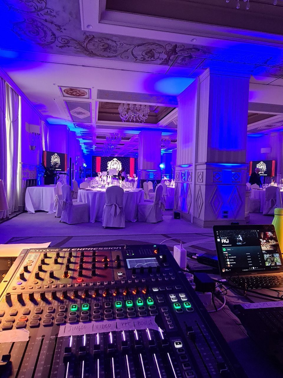 Attrezzatura per DJ con mixer e laptop, locale con illuminazione blu, tavoli rotondi e soffitto decorativo.