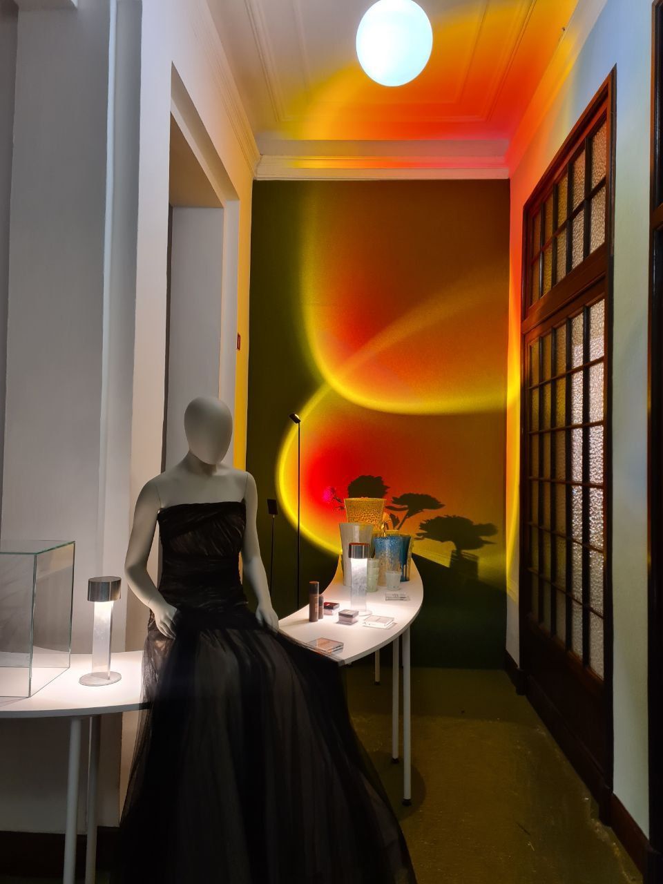 Manichino in abito nero, illuminato da una lampada effetto tramonto. Tavolo espositivo con oggetti, interno corridoio.