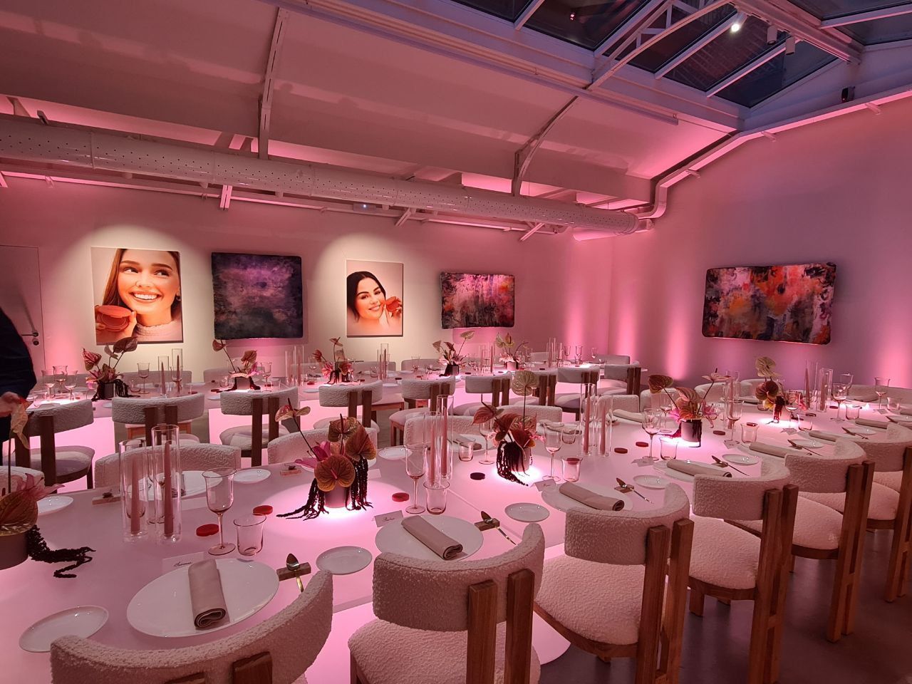 Uno spazio per eventi illuminato da luci rosa, con tavoli, sedie, centrotavola floreali e opere d'arte alle pareti.