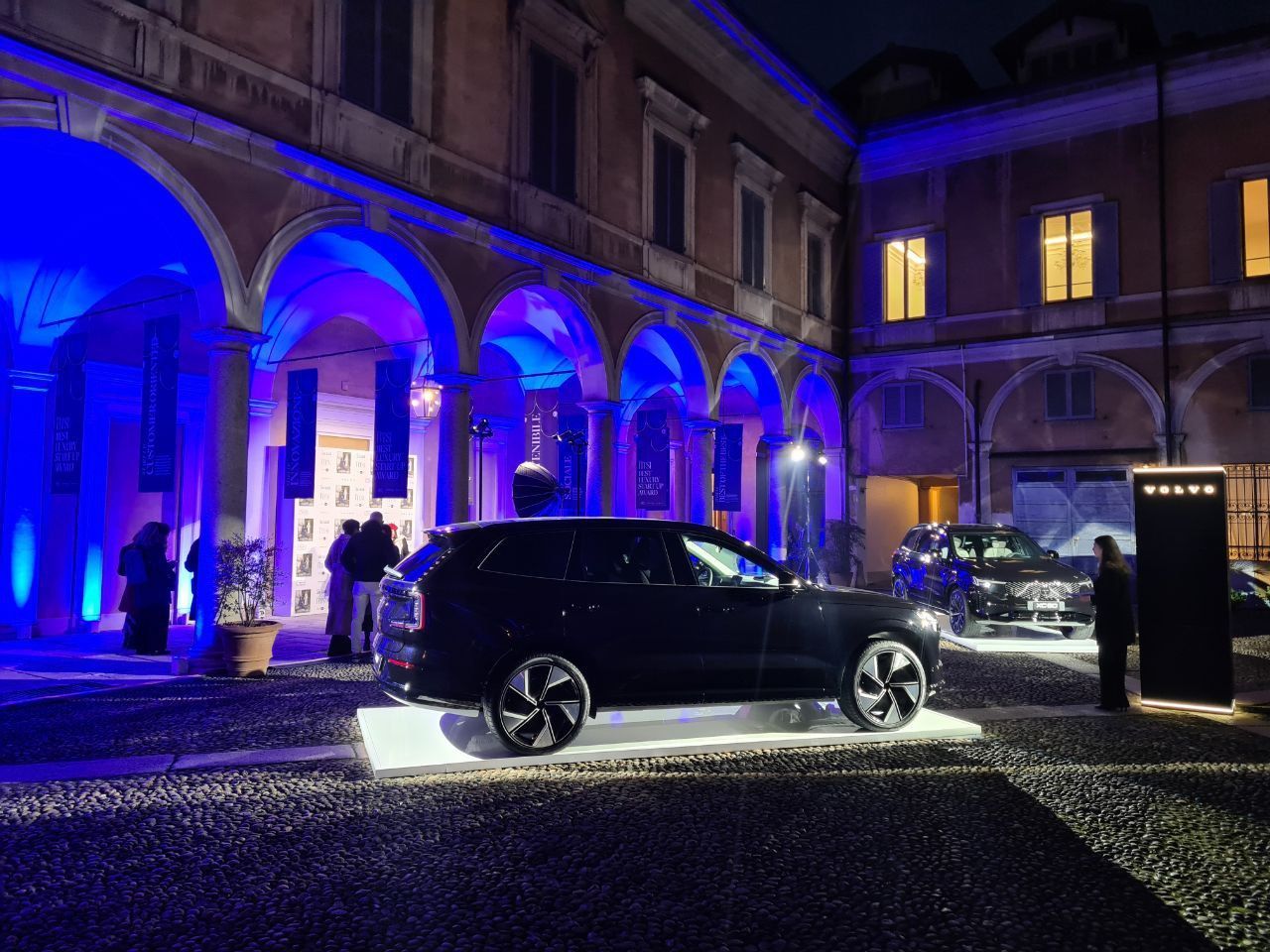 Due SUV scuri su piattaforme illuminate in un cortile, illuminati da luce blu, con gente che si aggira intorno.