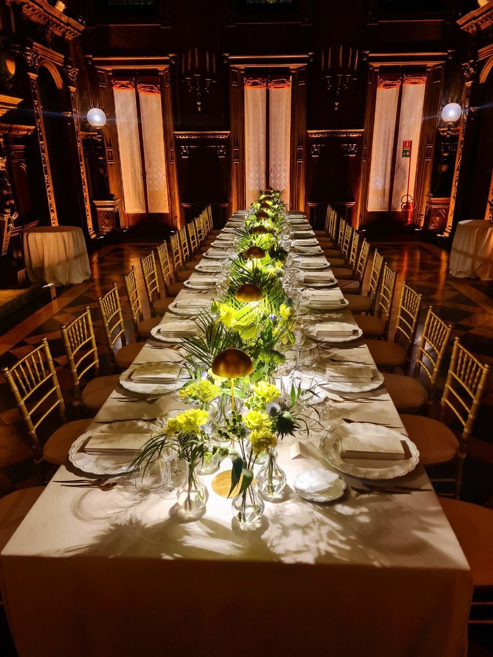 Lungo tavolo da pranzo apparecchiato per un evento formale, con centrotavola floreali, in un'ampia sala decorata.