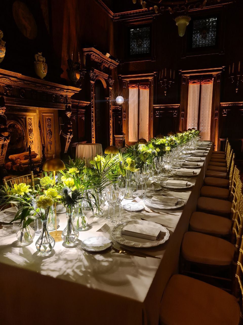 Lungo tavolo da pranzo apparecchiato per un evento formale, illuminato con toni caldi. Fiori gialli, bicchieri e sedie dorate.