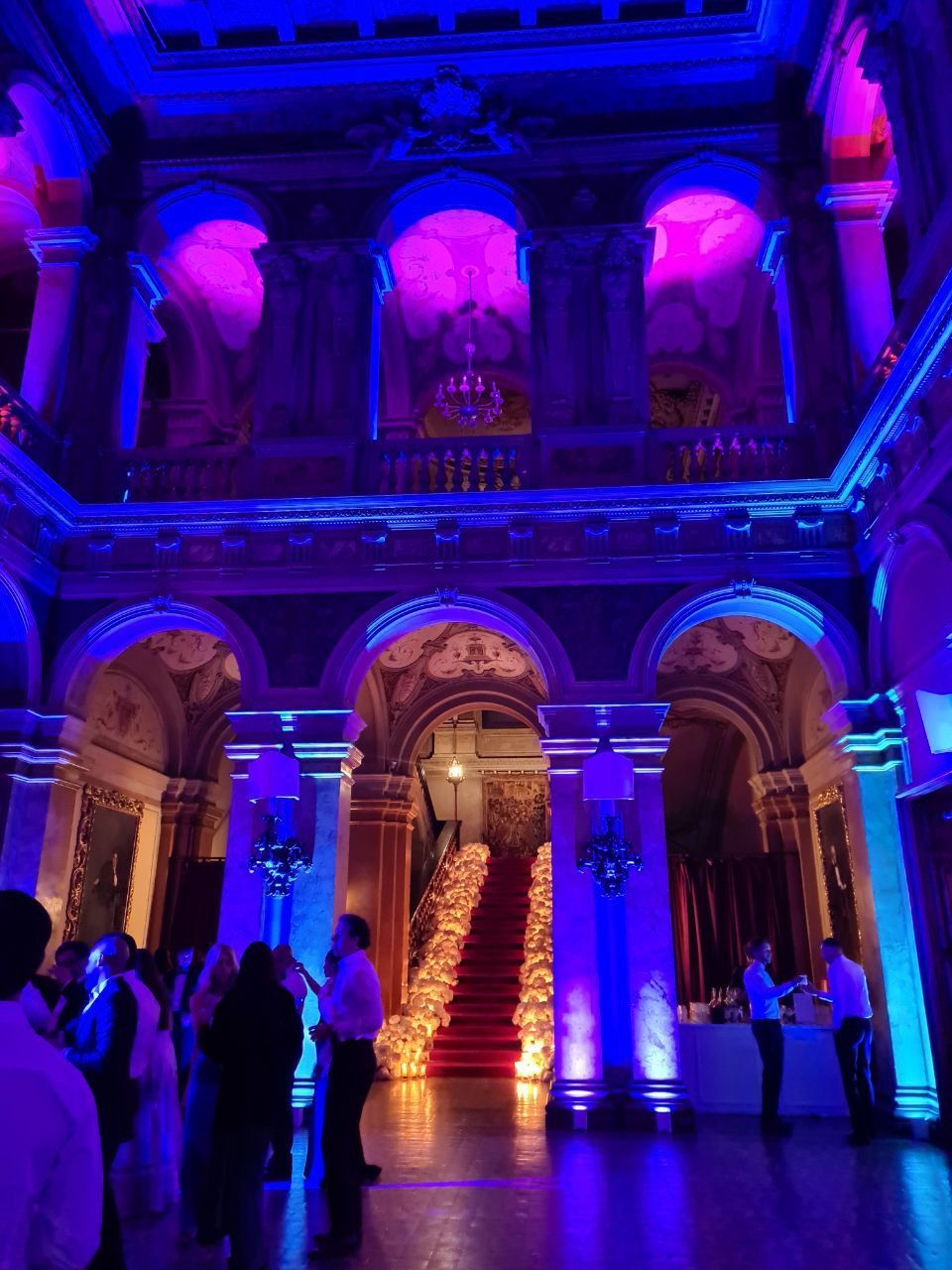 Una grande sala illuminata da luci viola e blu. La gente si raduna vicino a una scalinata ricoperta di moquette rossa.