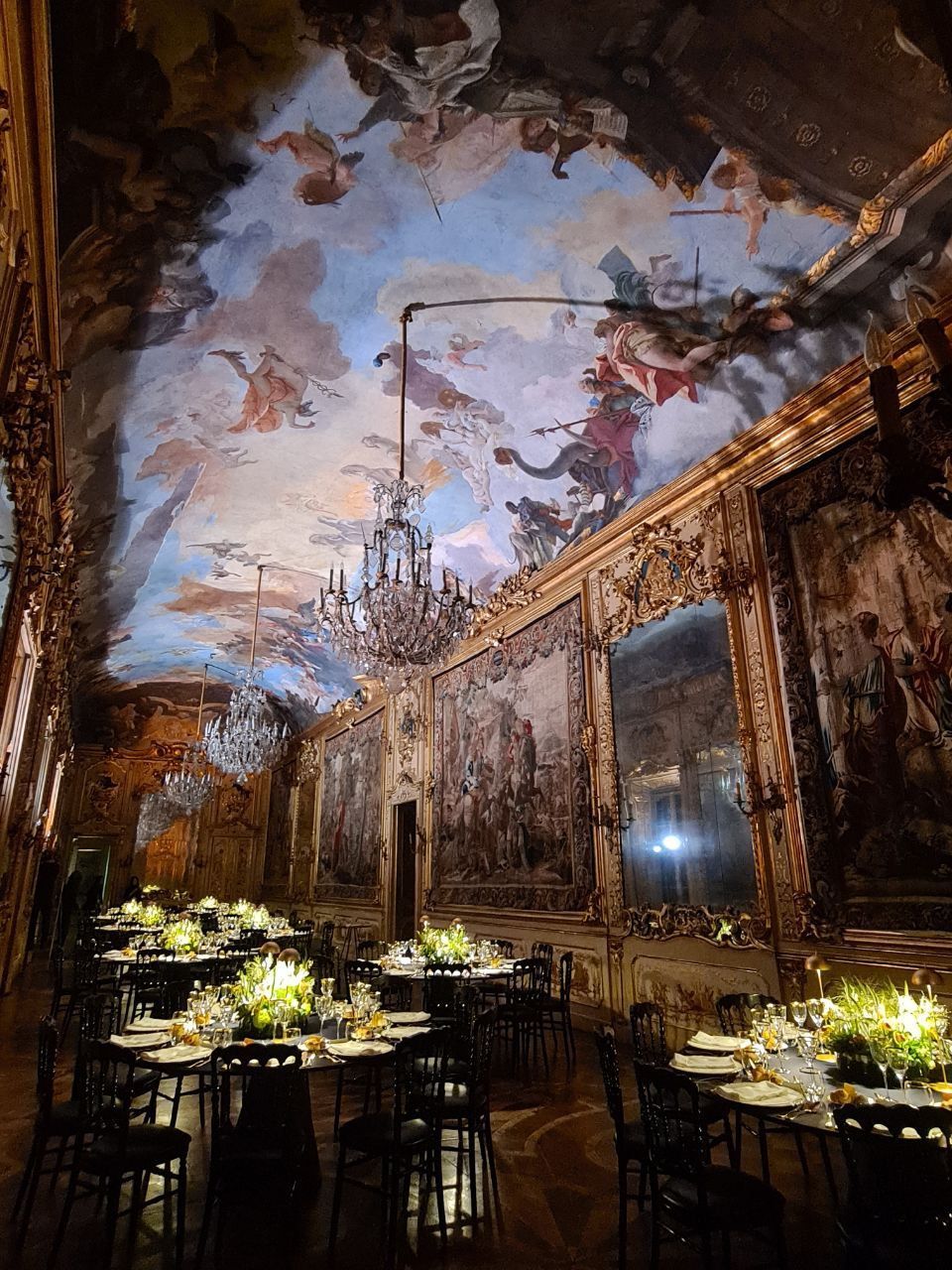 Stanza decorata con soffitto dipinto, lampadari, tavoli apparecchiati per cenare.