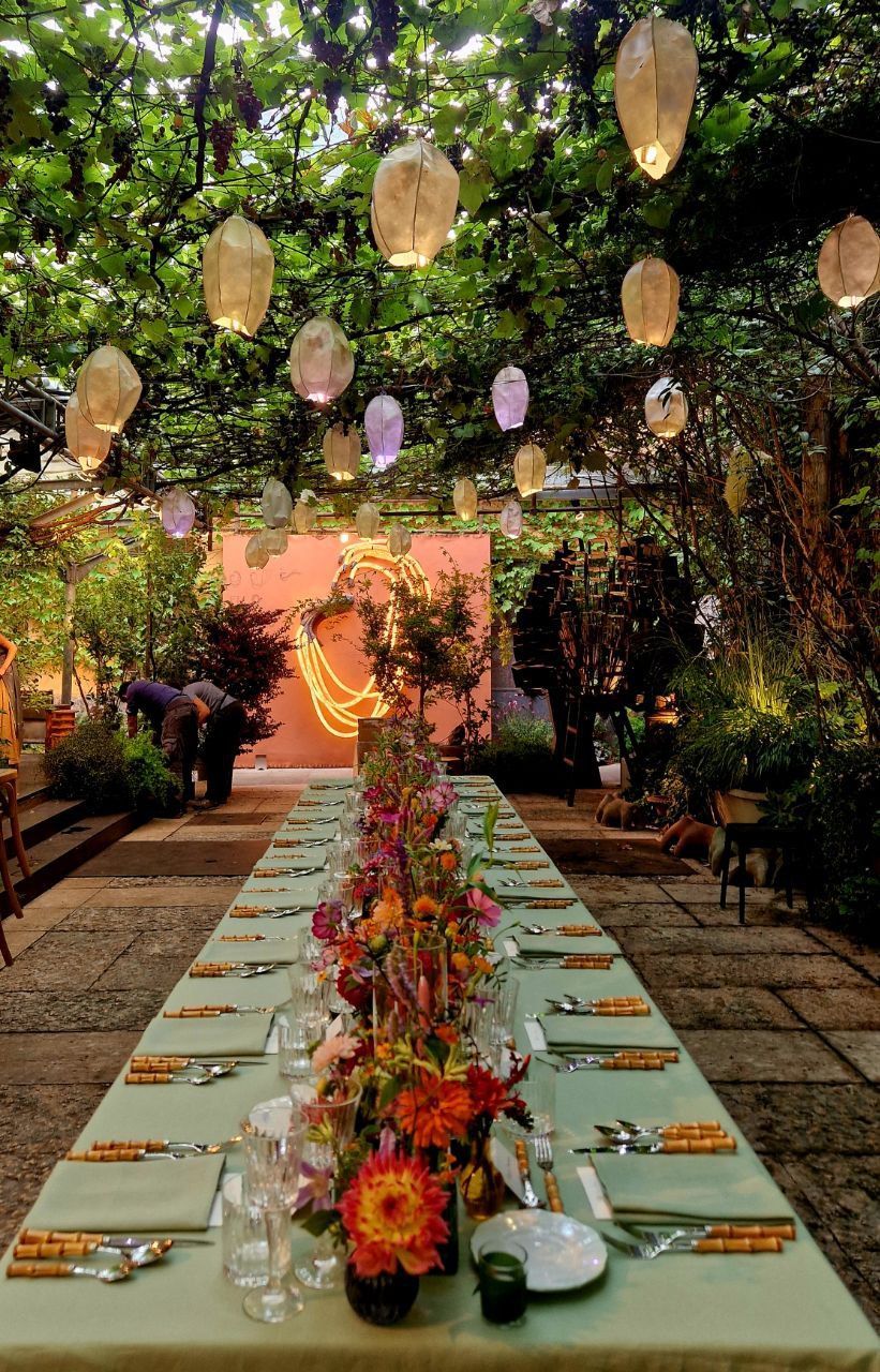 Lungo tavolo apparecchiato per un pasto, sotto viti e lanterne di carta in un giardino.