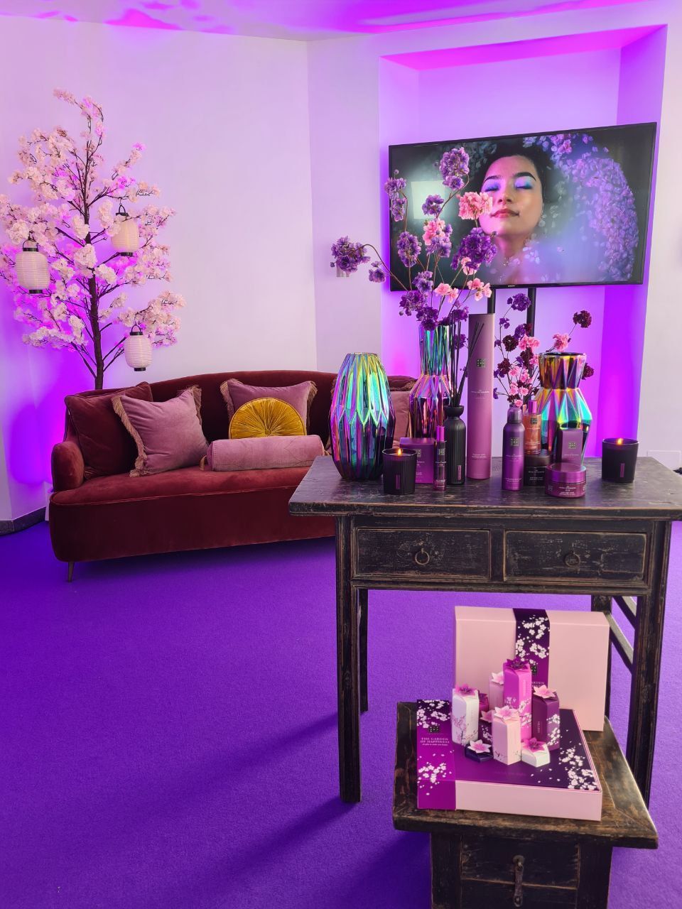 Stanza illuminata di viola con espositore per cosmetici, divano, fiori di ciliegio e una donna sullo schermo della TV.