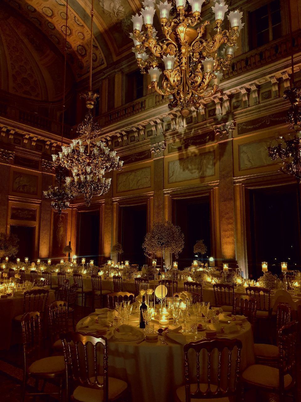 Elegante sala da ballo con tavoli rotondi apparecchiati per la cena, illuminata da lampadari e candele.
