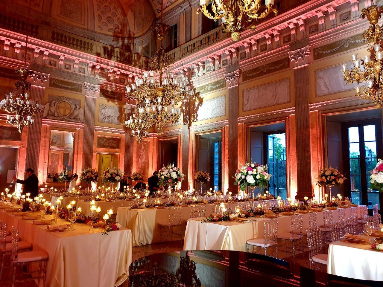 Elegante sala da ballo allestita per un evento formale con lunghi tavoli, centrotavola floreali, candele e lampadari; illuminazione rosa.
