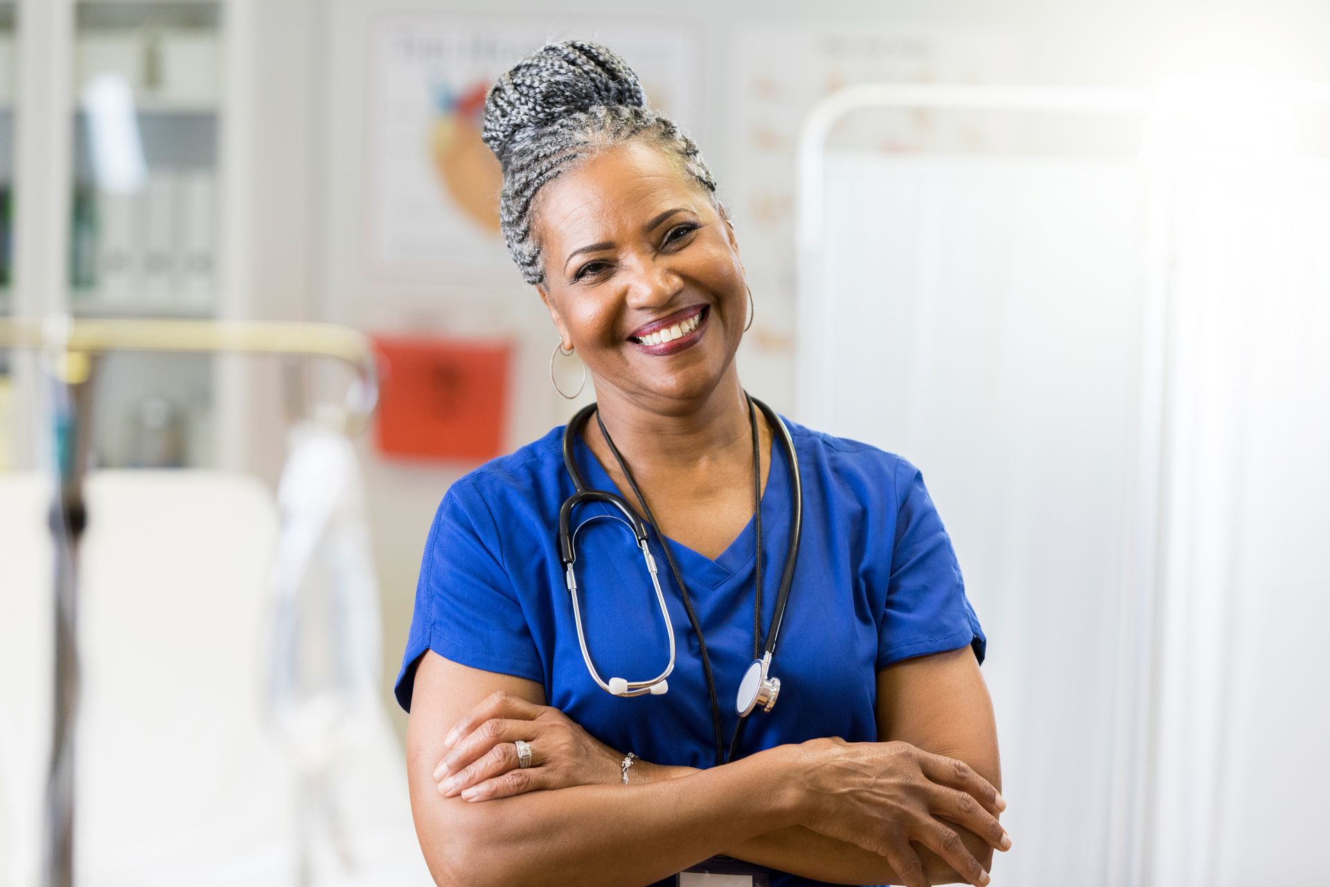 Smiling healthcare worker in blue scrubs with arms crossed, stethoscope, in a medical setting.
