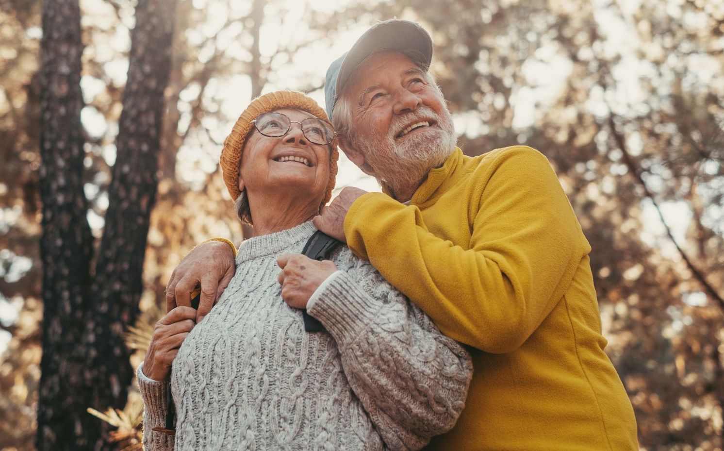 Senior couple smiles while looking up in a forest, man's arm around woman's shoulders, warm colors.