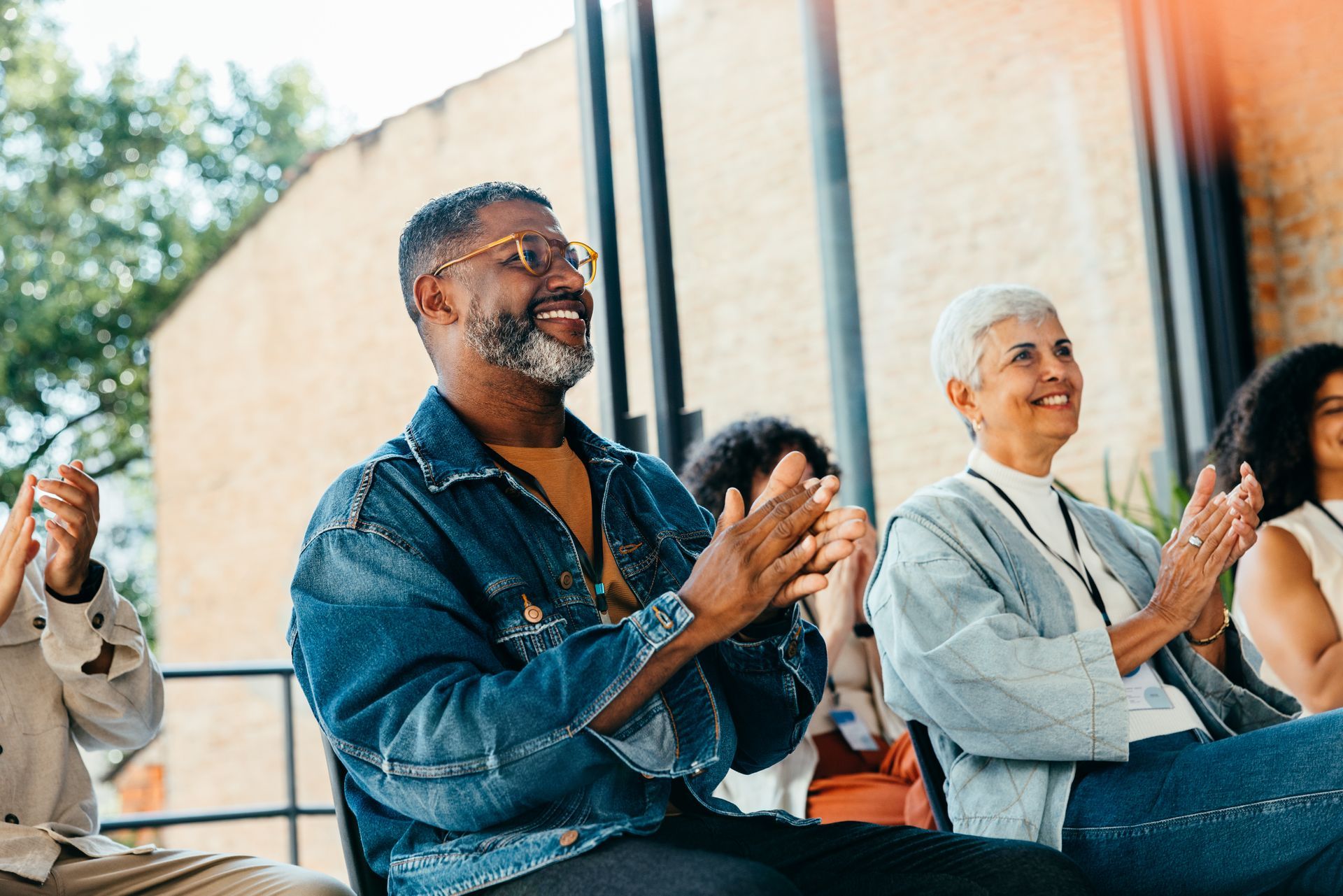 People clapping outdoors, smiling. Man in denim jacket, woman in light blue.