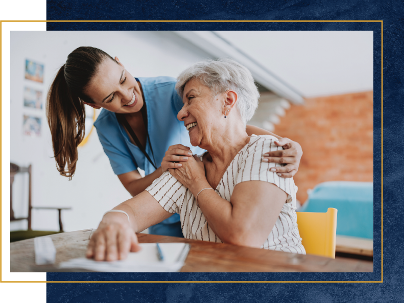 Caregiver smiles, comforting elderly woman at a table, both indoors.
