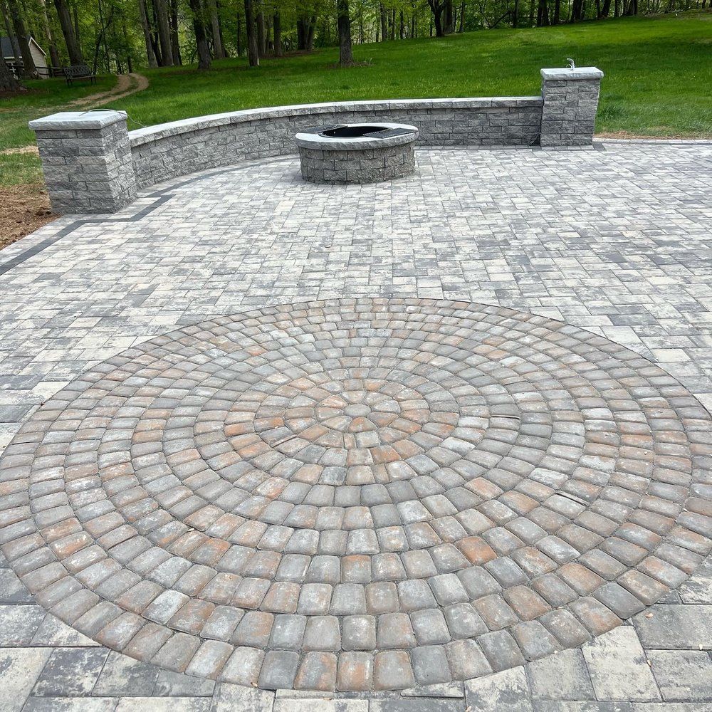 Paver patio with circular design, fire pit, and stone seating wall. Green lawn and trees in the background.