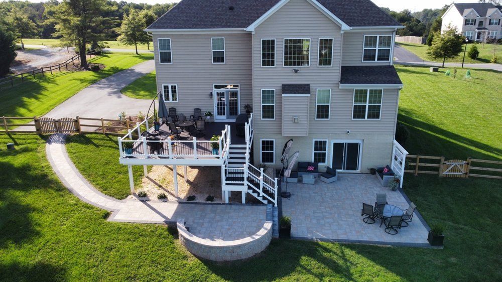 Back of a two-story beige house with a deck, patio, and walkway surrounded by green grass and a fence.