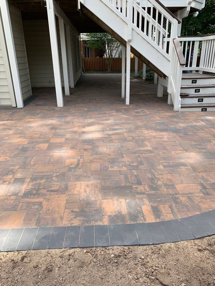 Brick patio under a deck with stairs, bordered by dark gray edging, tan siding.