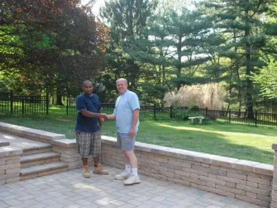 Two men shaking hands on a stone patio. One is Black, the other white. Green lawn, trees in the background.