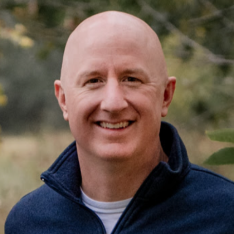 A bald man in a suit and tie is smiling in front of a brick wall.