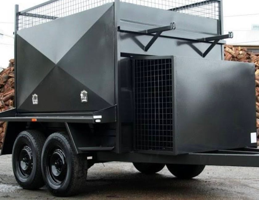 A black box trailer with tandem axels and a black box on top is parked in front of some wood.