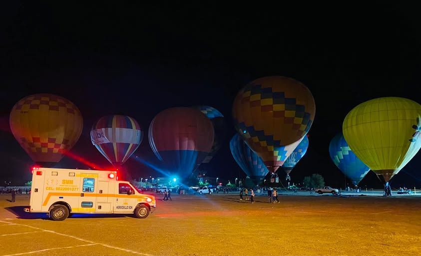 Una ambulancia blanca está estacionada frente a un campo de globos aerostáticos por la noche.
