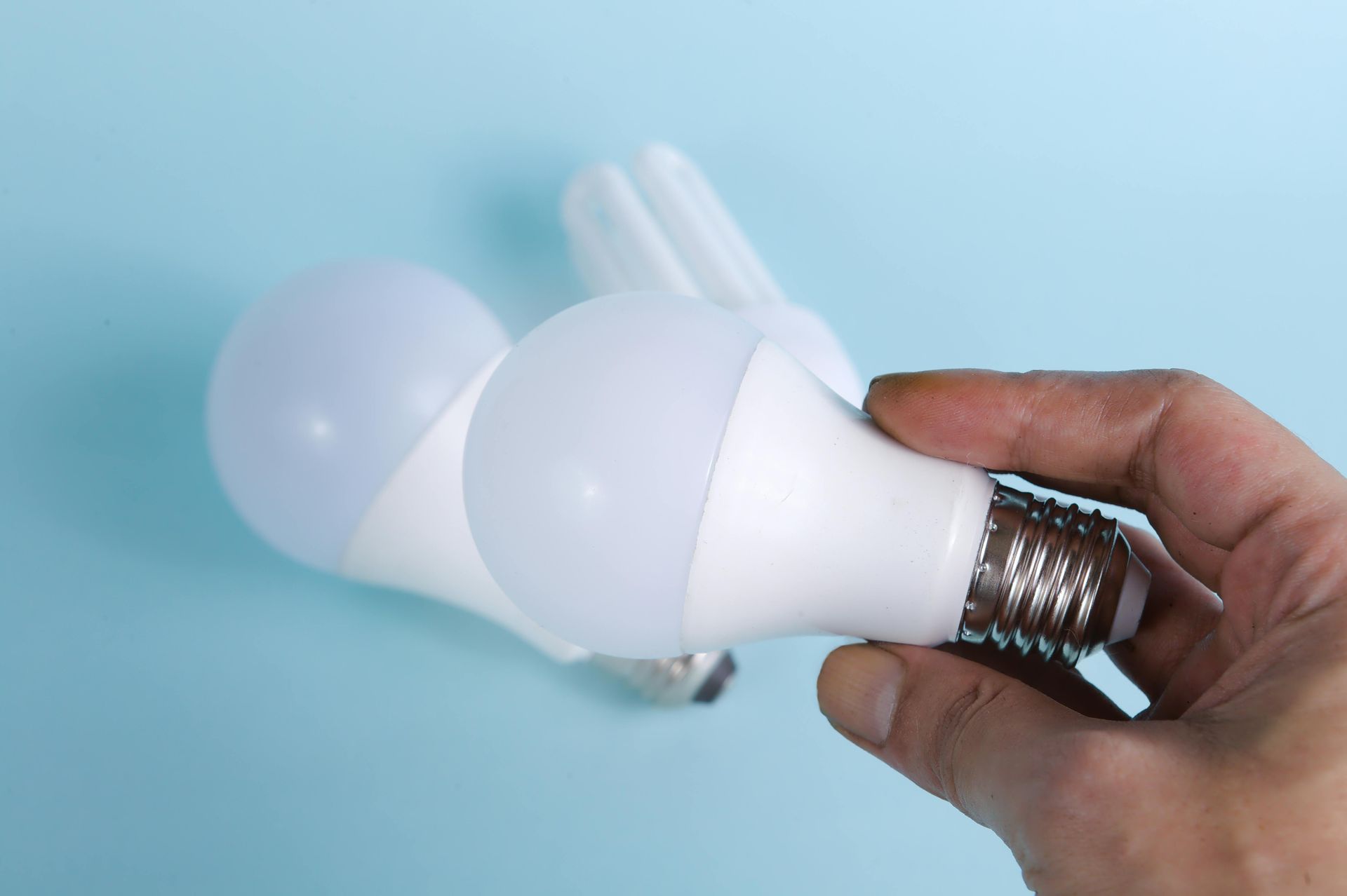 Hand holding a white LED lightbulb next to two others, against a blue background.