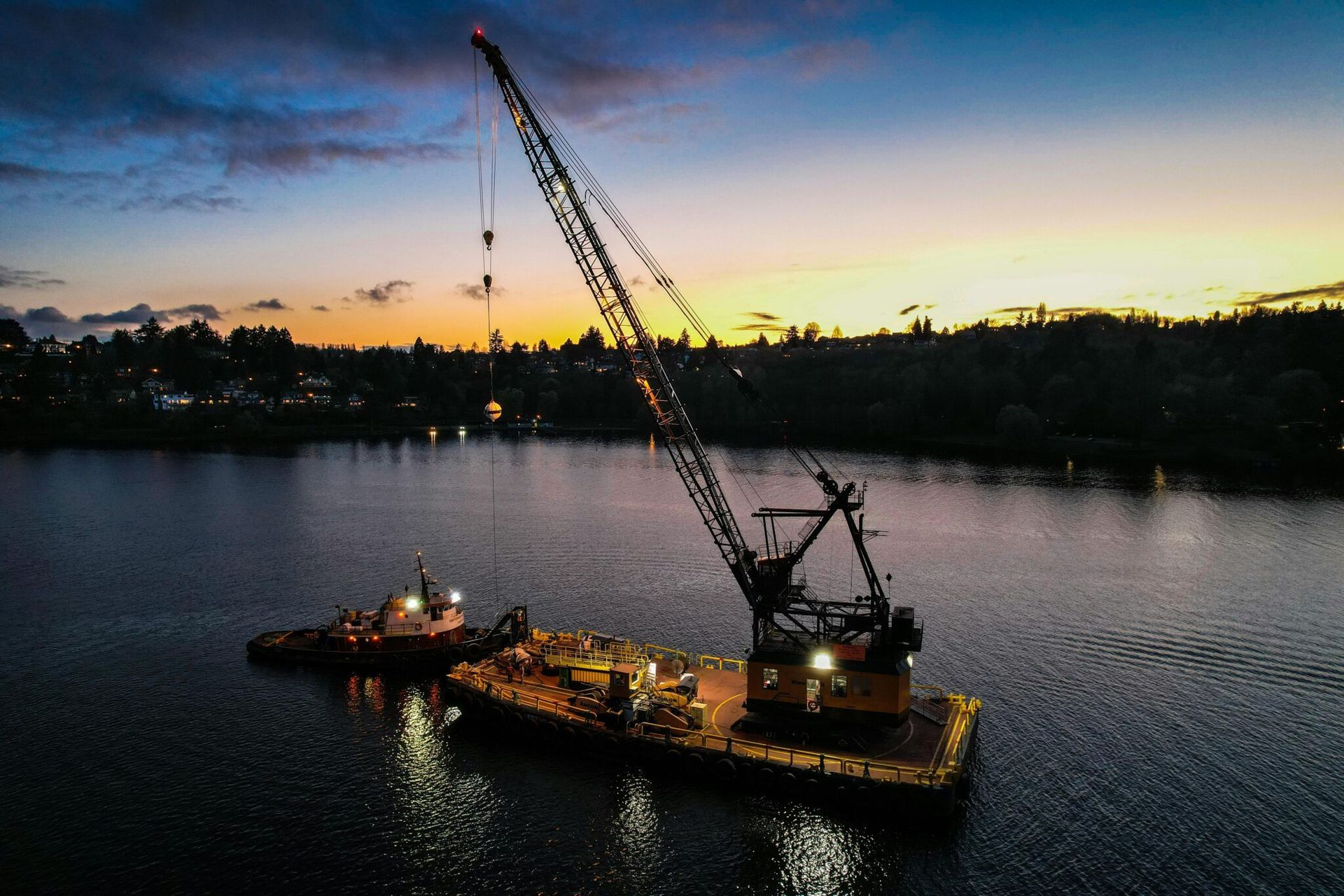 Crane barge on a calm river at dusk with city lights and a colorful sunset sky