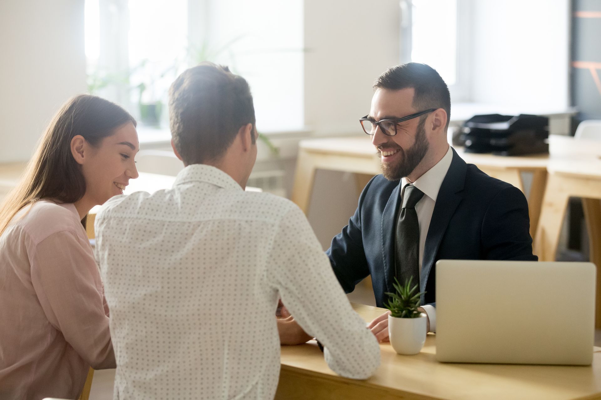 Three people in a bright office meeting, talking across a desk with a laptop and coffee cup.