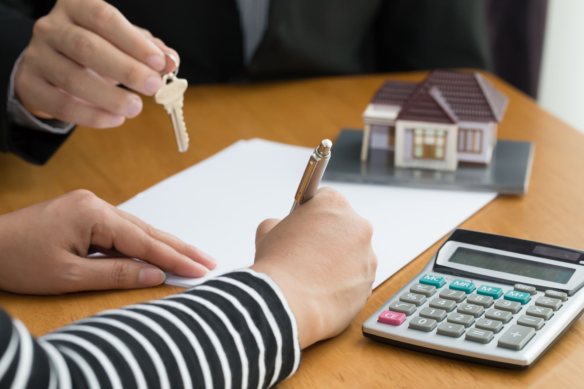 Hands exchanging house keys over a desk with a calculator and house model