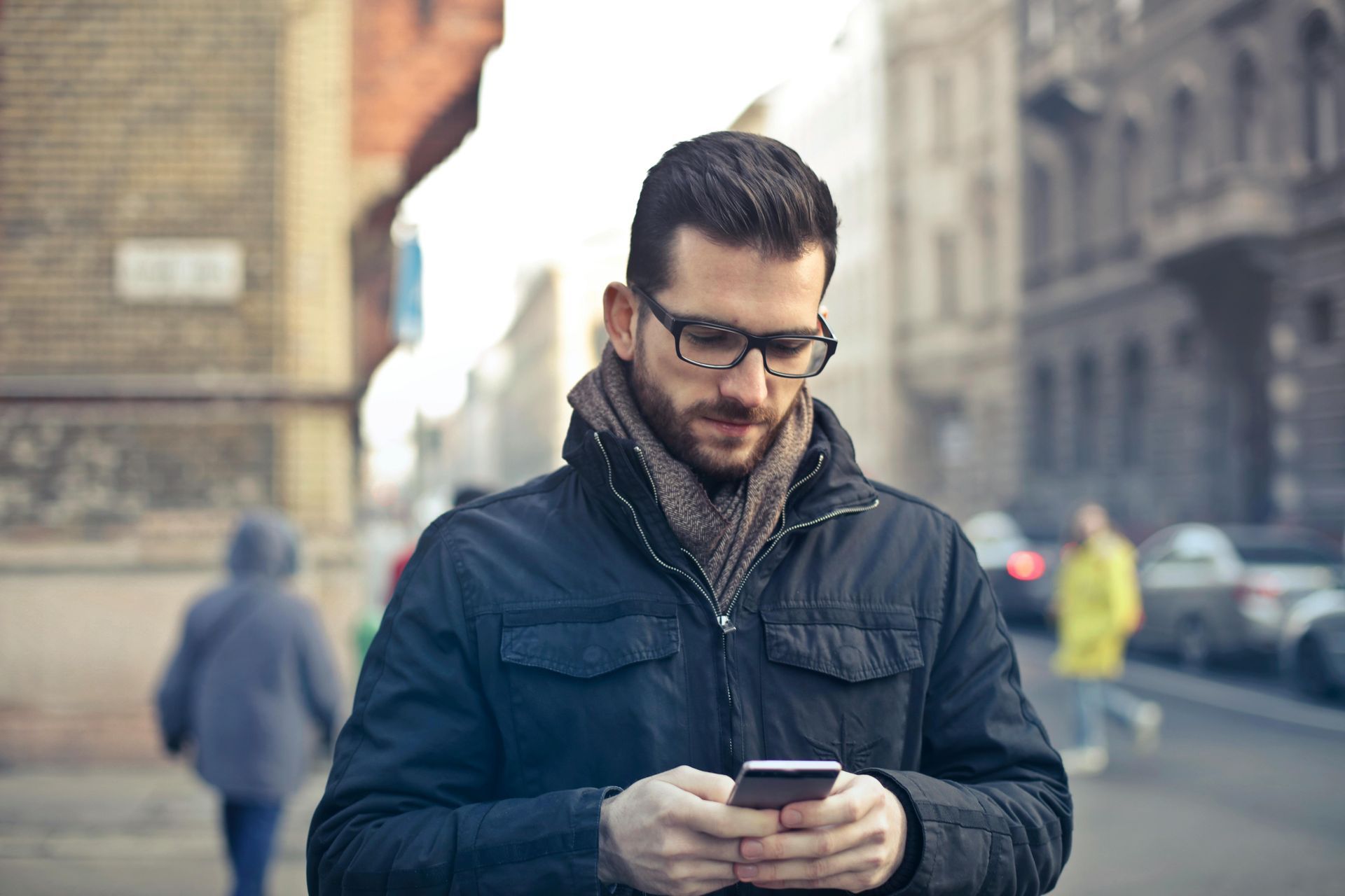 Man in glasses and scarf looking at his phone on a city street.