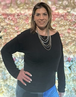 Woman in black top and multi-strand necklace, hand on hip, in front of flower wall.