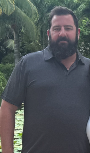 Man with a full beard wearing a gray polo shirt, standing outdoors in front of green foliage.