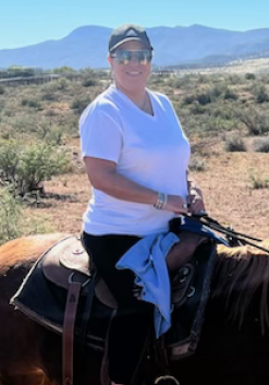 Woman riding a horse in a desert landscape; wearing a white shirt, black pants, and a baseball cap.