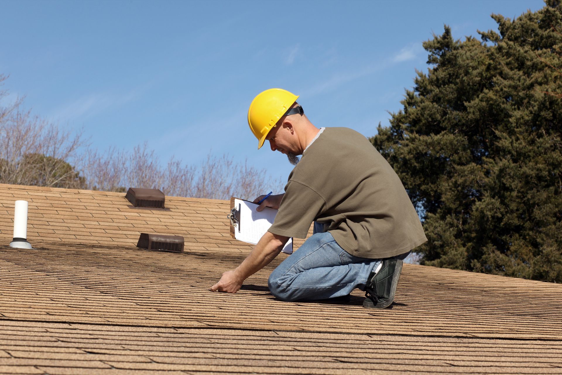 Worker in a yellow hard hat inspects a shingled roof with a clipboard.