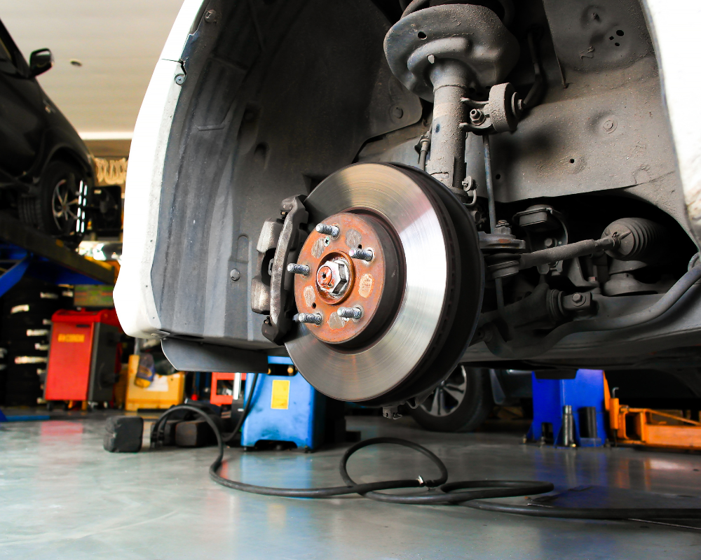 Car's front wheel removed, revealing brake rotor, caliper, suspension components, and jack stands in a garage.