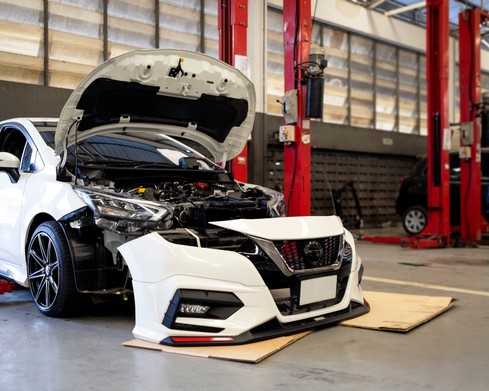 White car in a repair shop with hood open and front bumper removed, revealing the engine.
