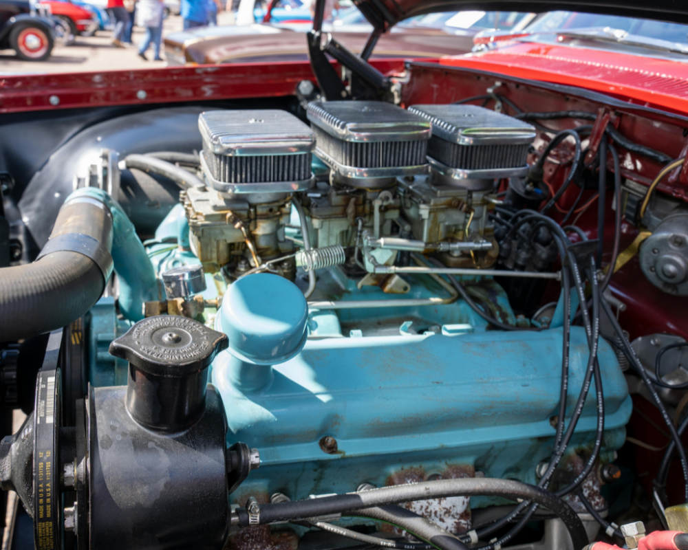 Red classic car engine with three carburetors, turquoise block, and black accessories, outdoors.