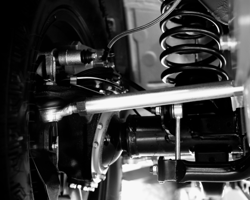 Black and white close-up of a vehicle's suspension system, including coil spring, control arm, and axle.
